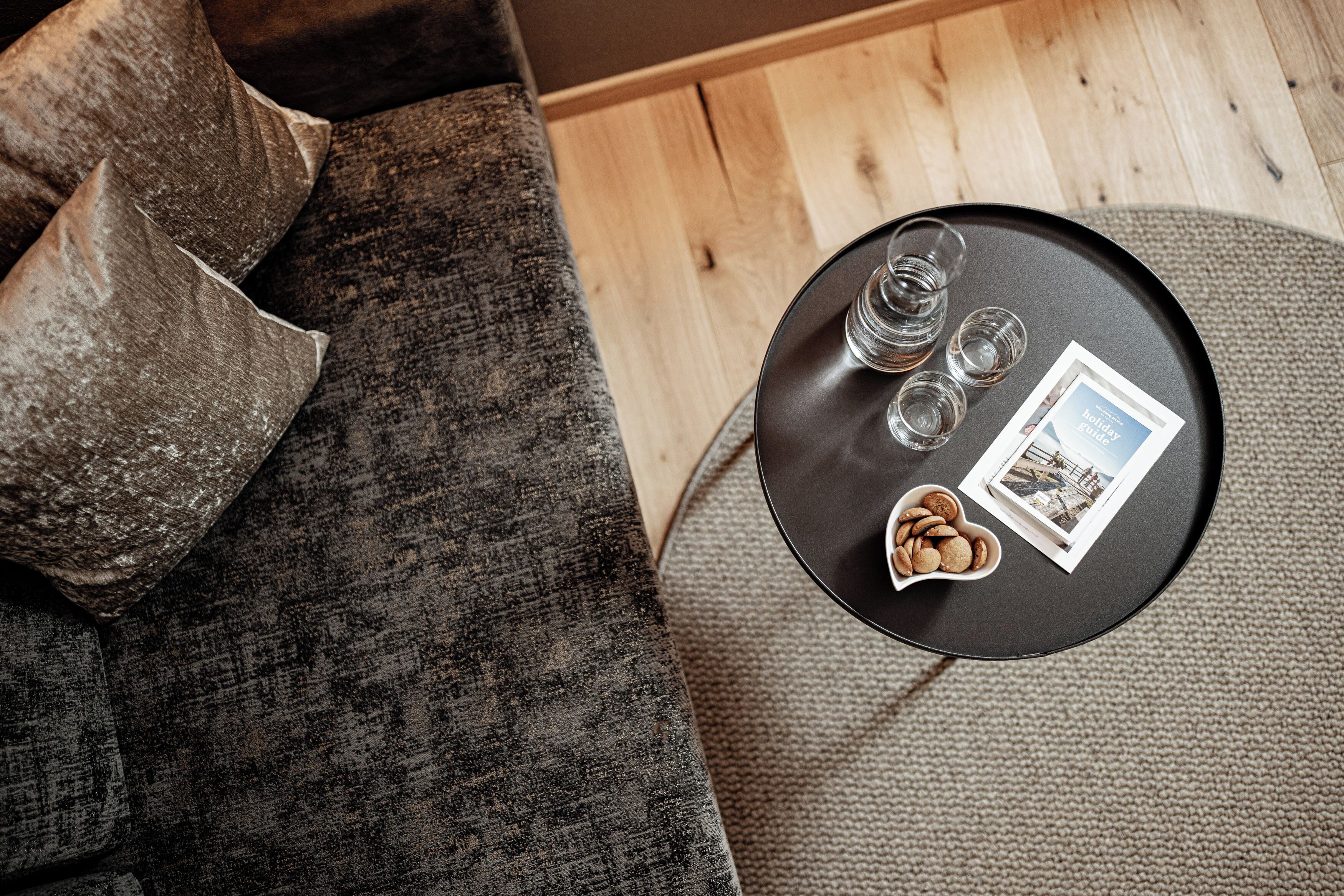 A round black table with a brochure, three glasses, and a bowl of cookies on a grey couch.