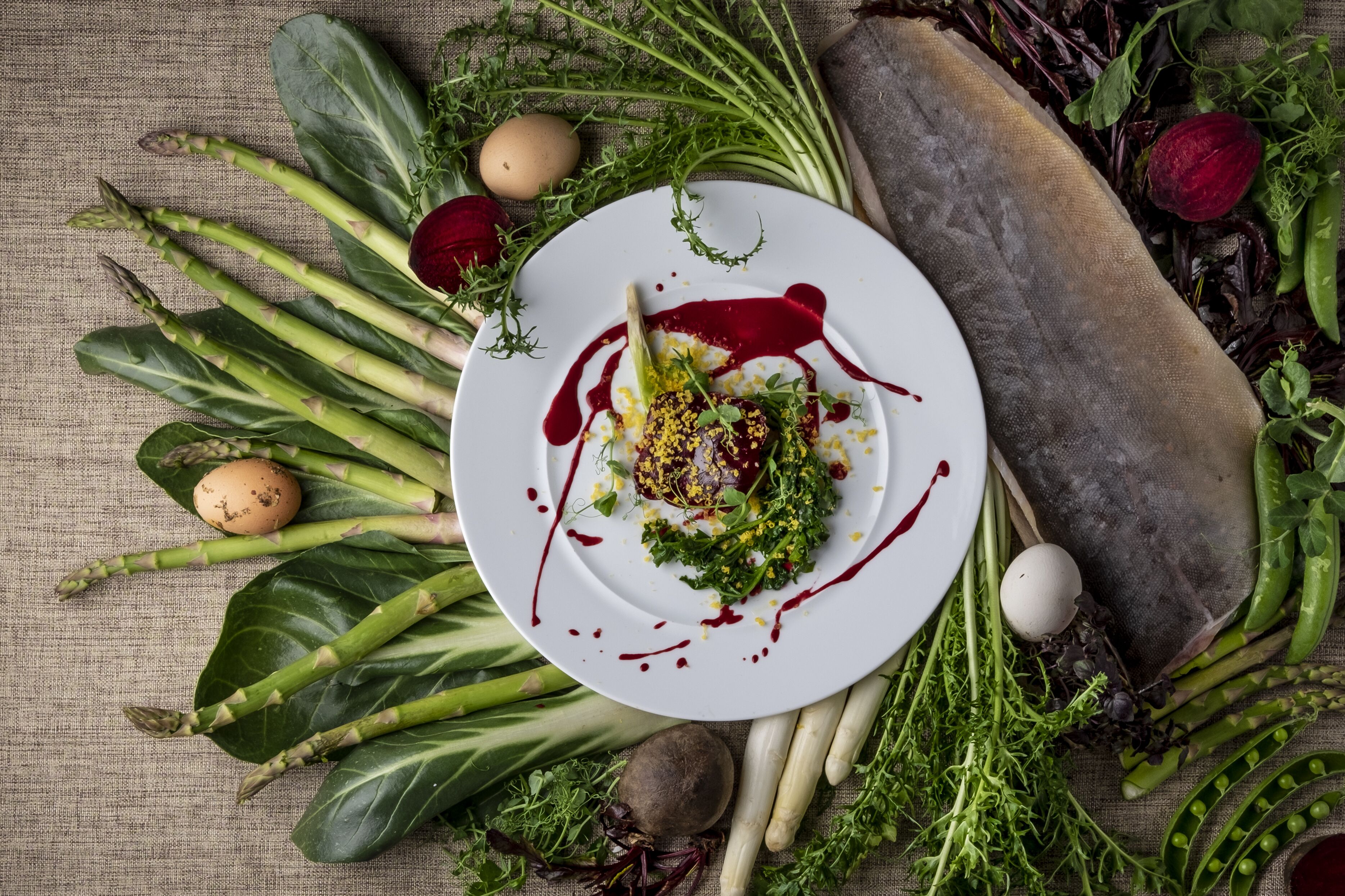 Un plato de comida con verduras y una remolacha, rodeado de más verduras y huevos sobre una superficie texturizada.