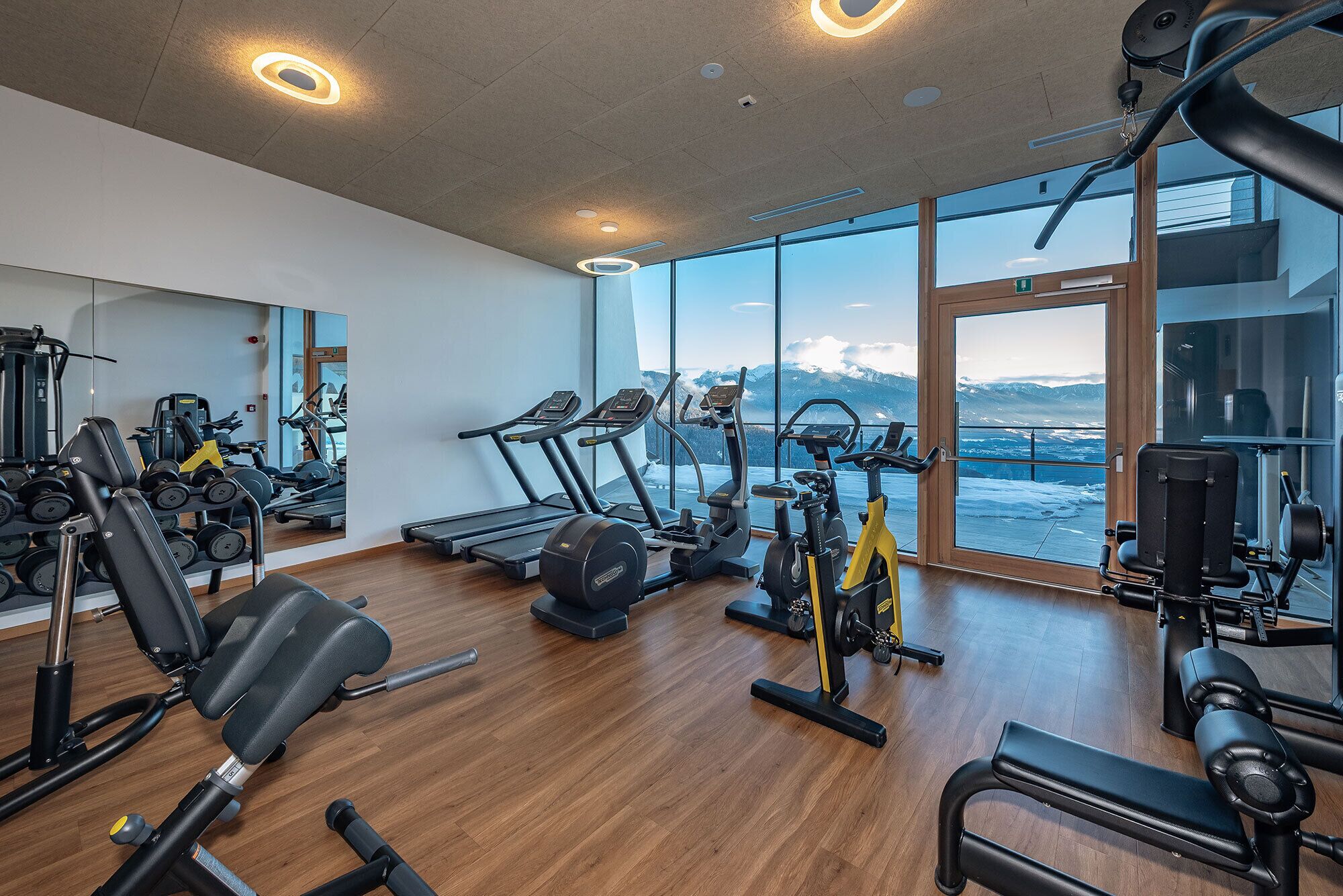 Un gimnasio moderno con máquinas de cardio, equipo de levantamiento de pesas y un amplio ventanal con vistas a la montaña.