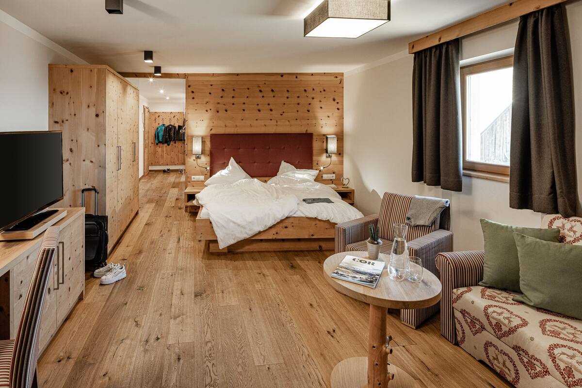 Une chambre d’hôtel avec un plancher en bois, un lit avec une literie blanche, une télévision, un canapé, une petite table et une fenêtre avec des rideaux.
