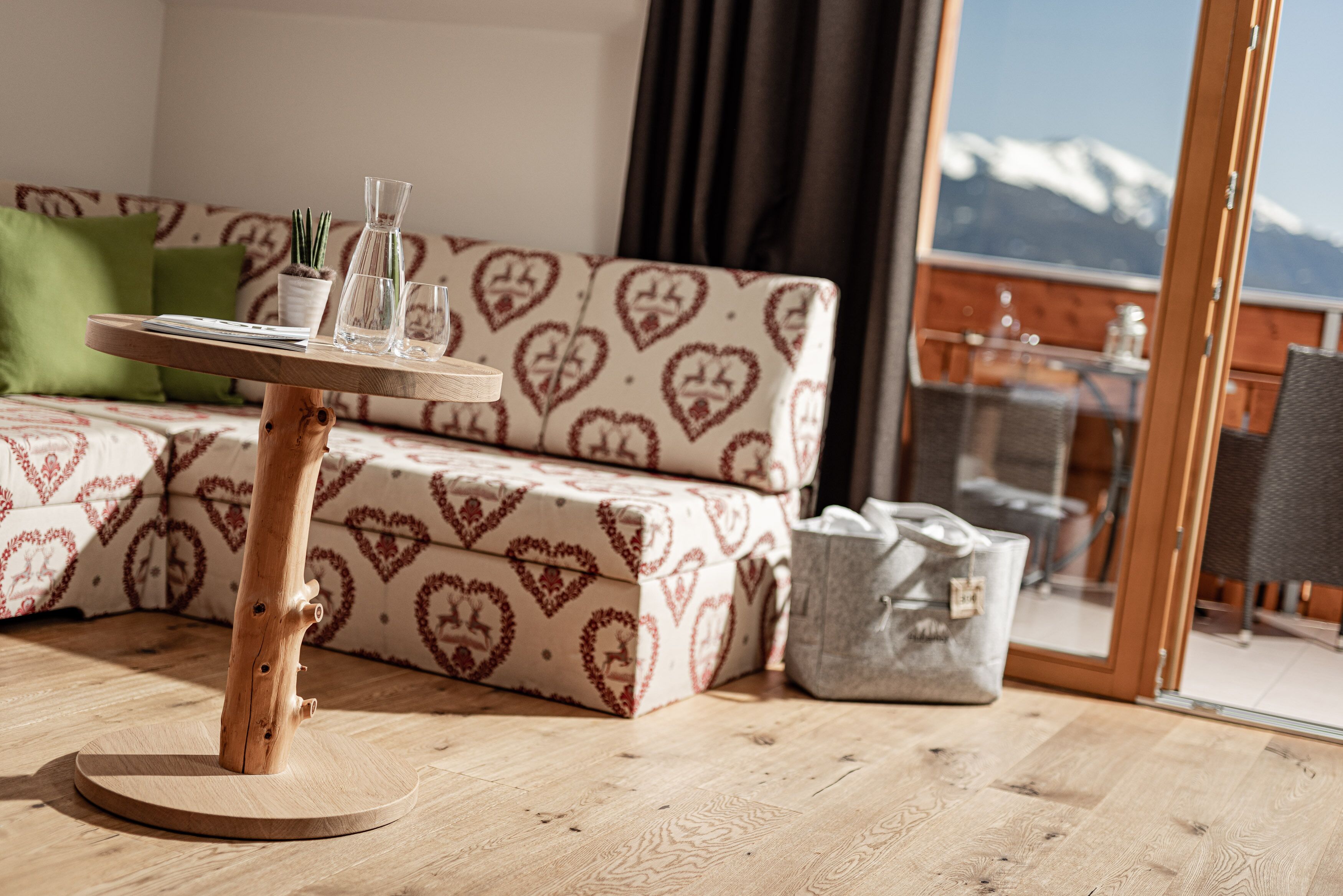 A cozy room with a patterned sofa, a wooden coffee table with glasses and a plant, and a view of mountains through a glass door.