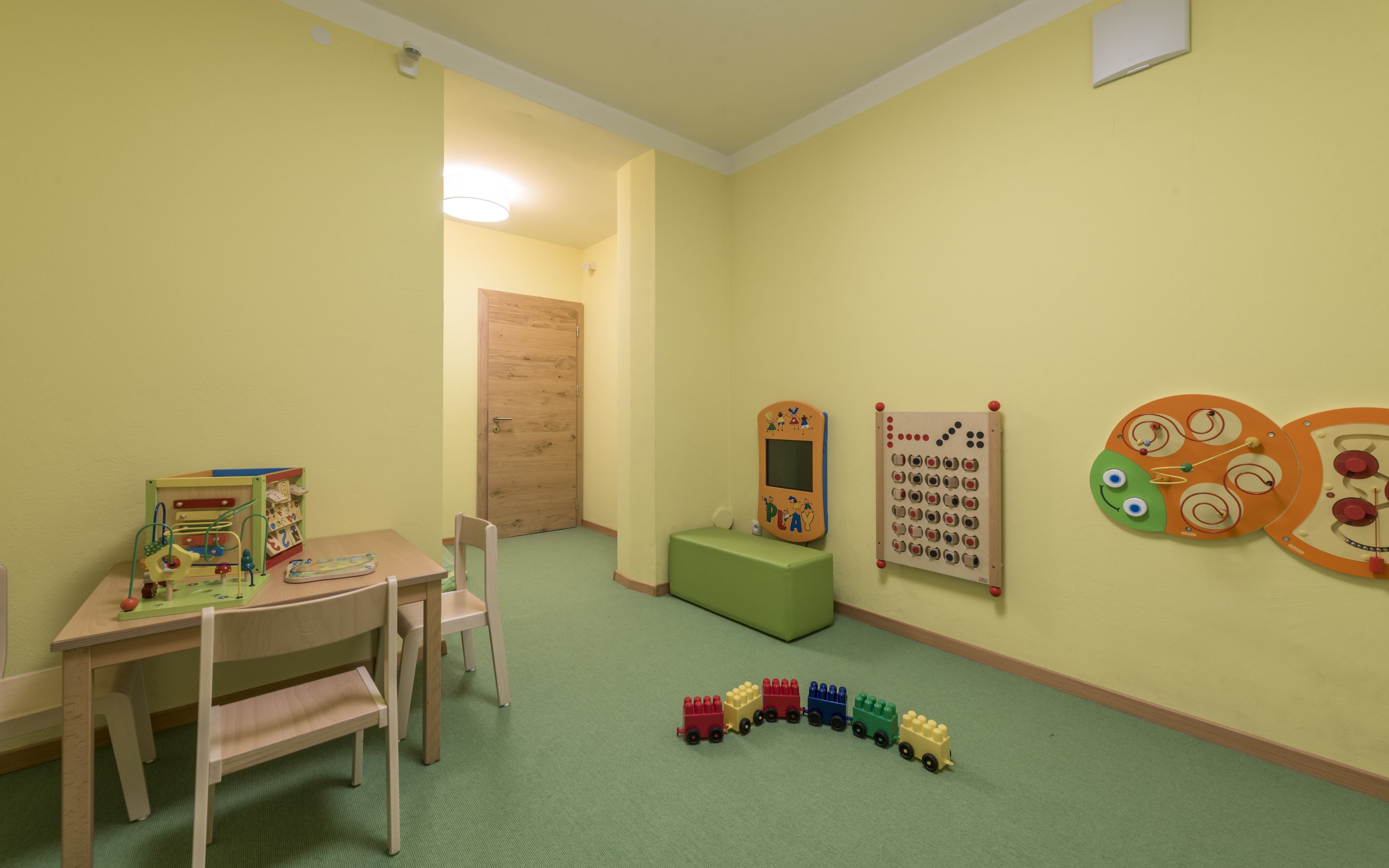 Una sala de juegos con una mesa de madera, sillas, un tapete de juego verde, un tren de juguete y adornos educativos en la pared.