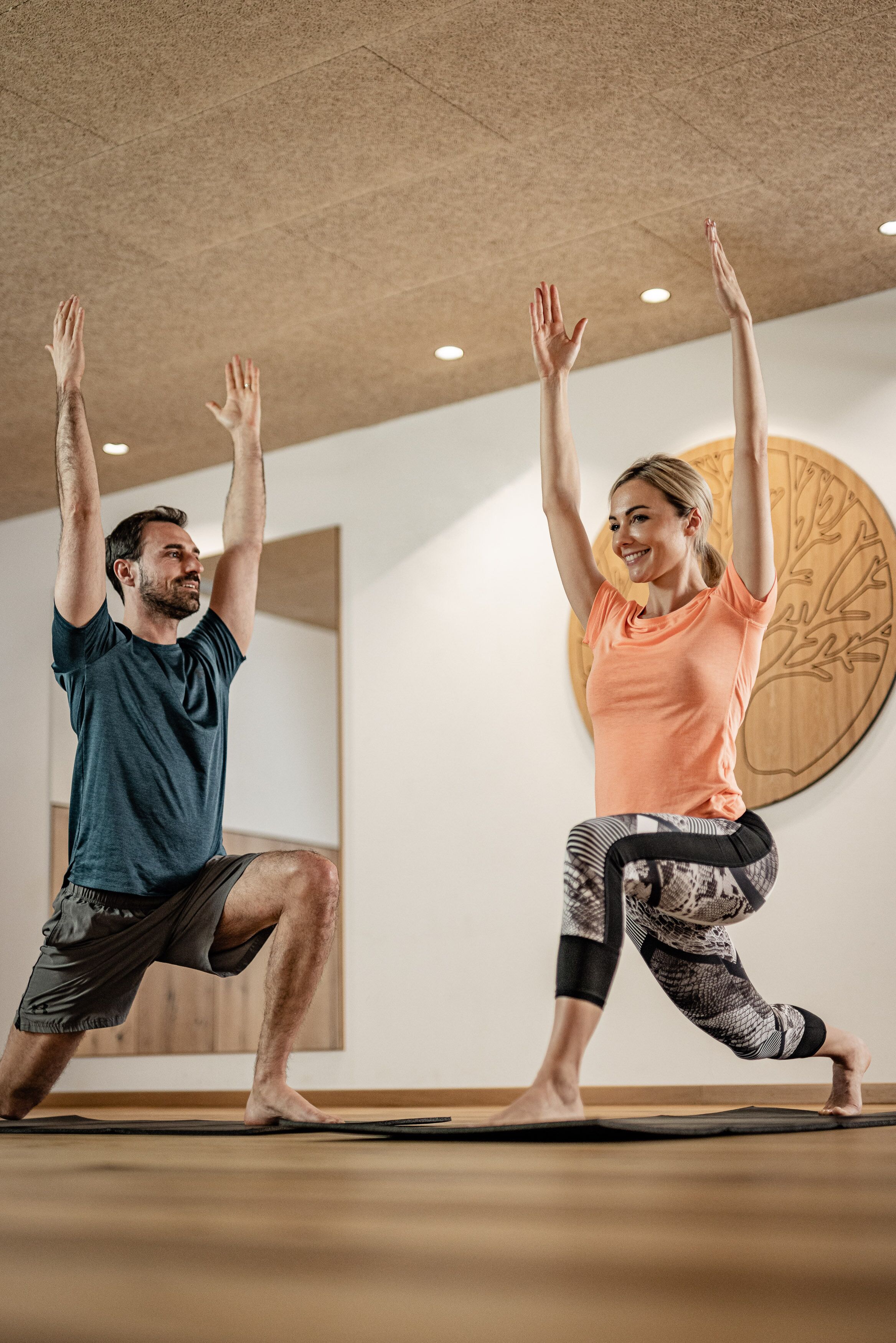 Two people performing a yoga pose in a room with a wooden ceiling and a large circular wall decoration.