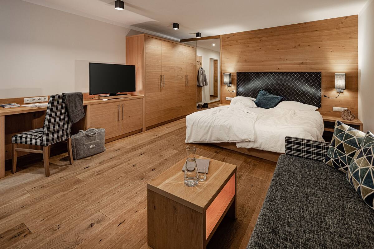 Une chambre d’hôtel moderne avec un plancher en bois, un lit avec une literie blanche, un canapé gris, un bureau en bois avec une chaise, une télévision et une armoire.