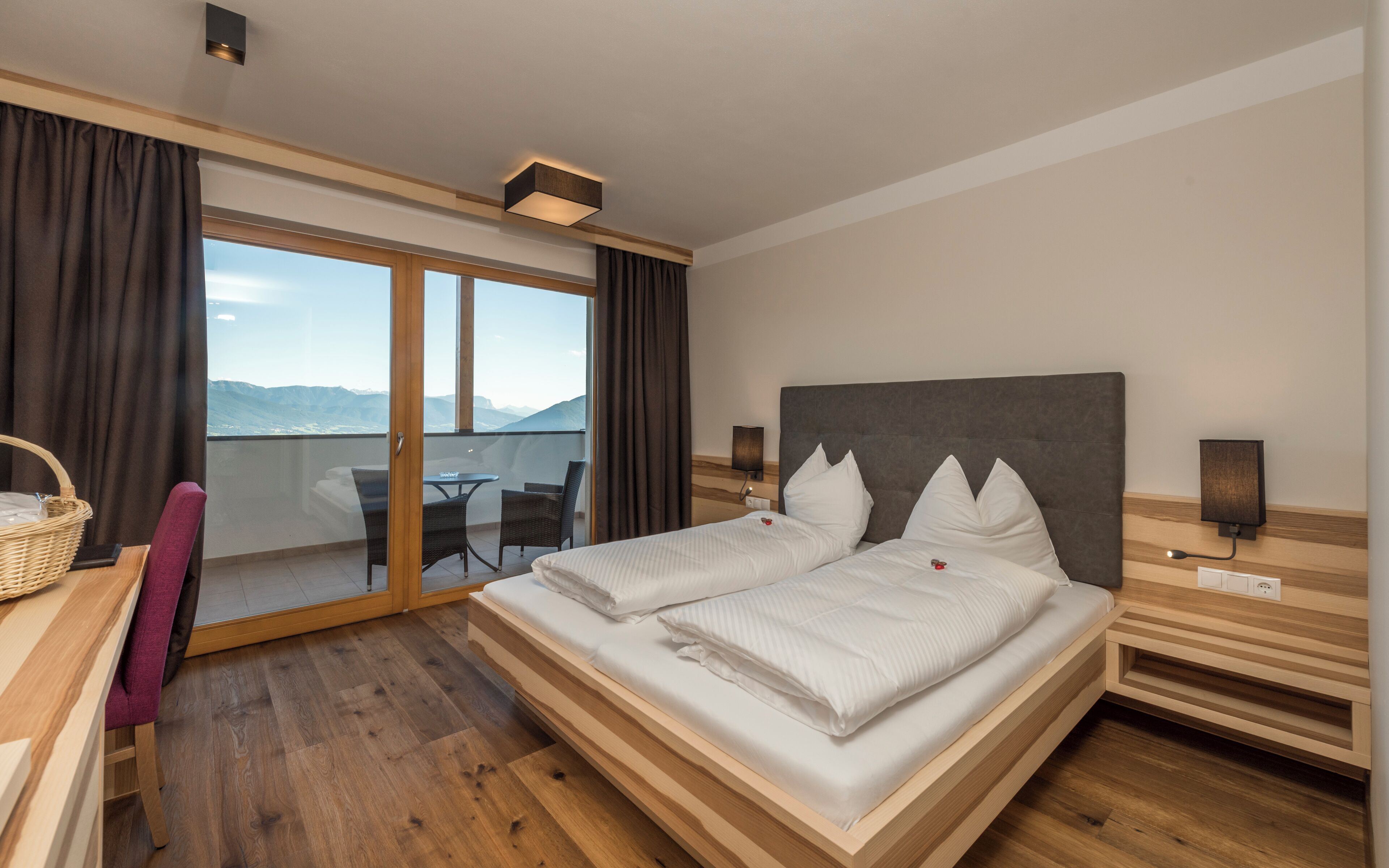 A hotel room with a large bed, a balcony with a view, wooden flooring, and a desk with a chair.