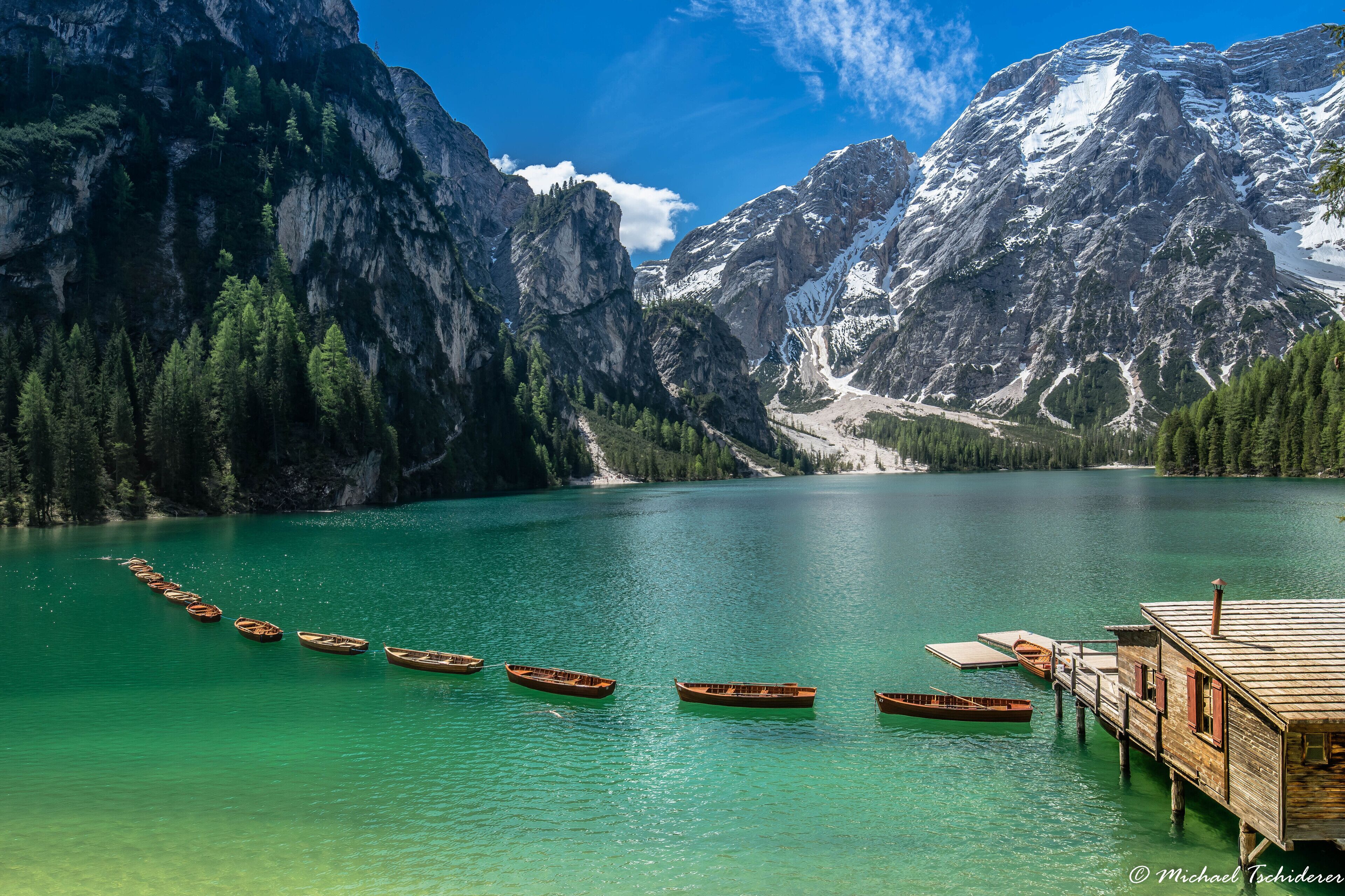 A wooden dock with several boats lined up on a turquoise lake, surrounded by forested mountains.
