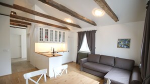 Un espacio de sala acogedor con una mesa de madera, sillas blancas, un sofá marrón y una cocina con armarios blancos y un ventanal con cortinas.