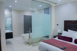 A modern hotel room with a glass-enclosed shower, a large bed with a red blanket, a bedside table with a lamp, and a nightstand with a remote control. - L'eola Suites (Abuja)
