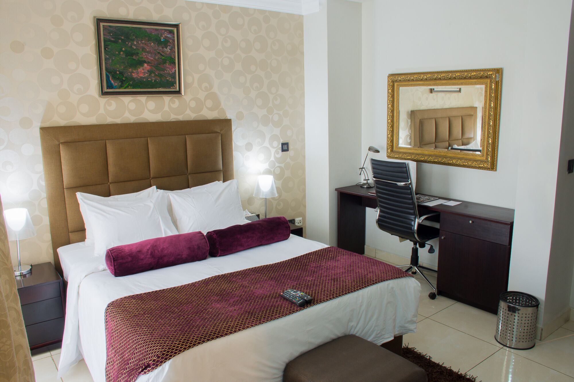 Una habitación de hotel con cama, escritorio, silla, lámpara, espejo y un cubo de basura.