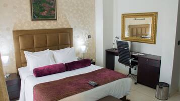 Una habitación de hotel con cama, escritorio, silla, lámpara, espejo y un cubo de basura.