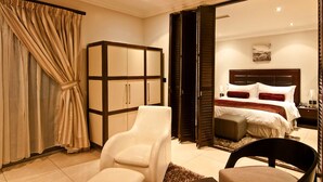 A bedroom with a large bed, a wardrobe, a sitting area with a chair, and a view through an open door. - L'eola Suites (Abuja)
