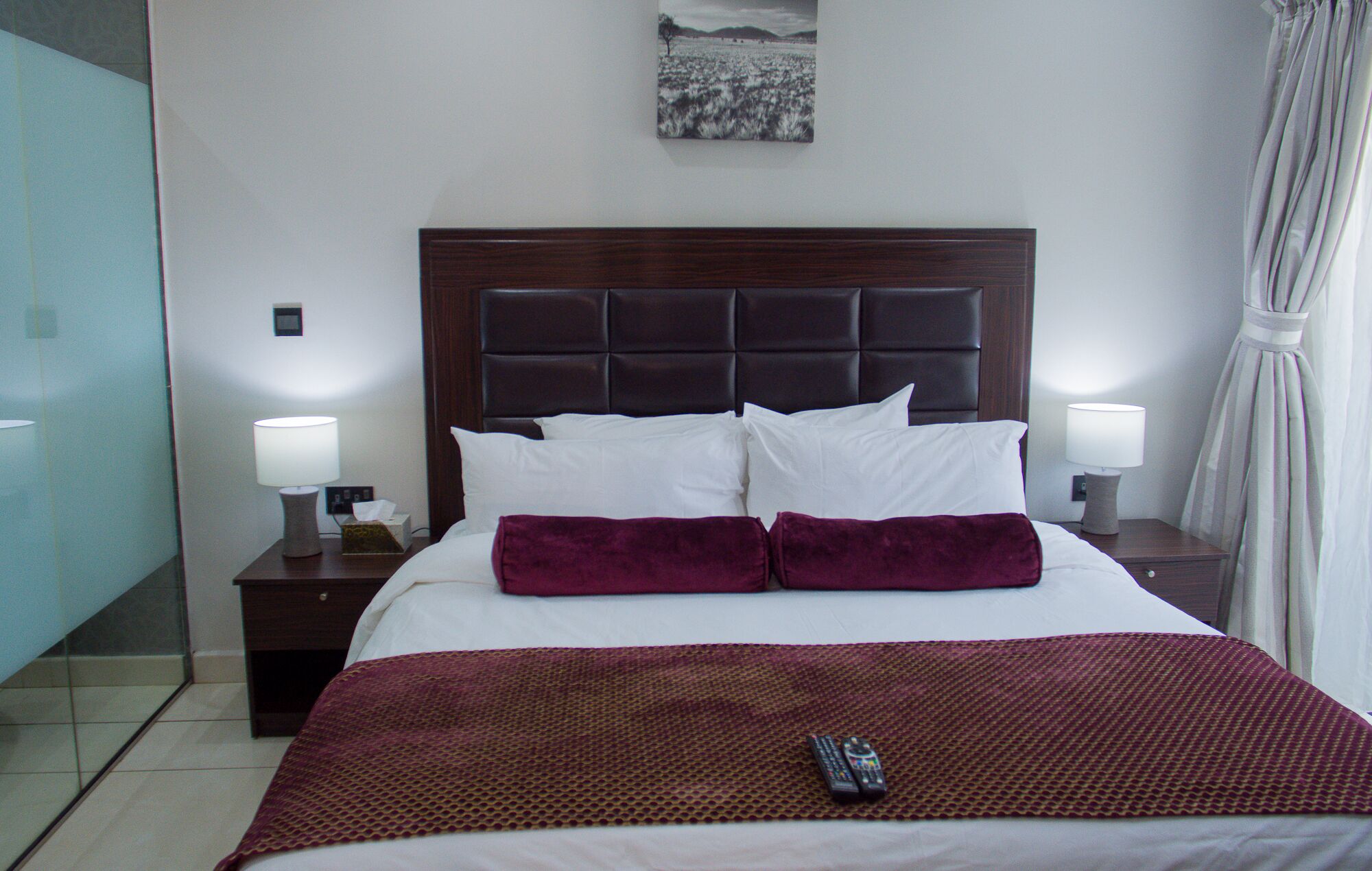A hotel room with a large bed, two bedside tables, a nightstand, a remote control, and a painting on the wall.