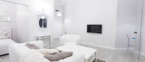 A modern, white-themed bedroom with a bed, two white sofas, a small table, a TV, and a chandelier.