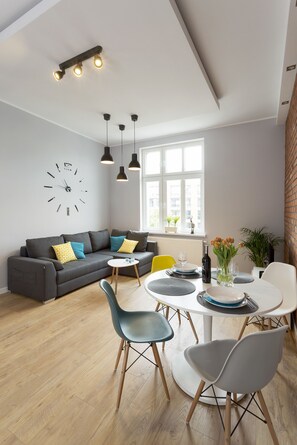 A modern living room with a grey sofa, a dining table with chairs, a large window, and a wall clock.