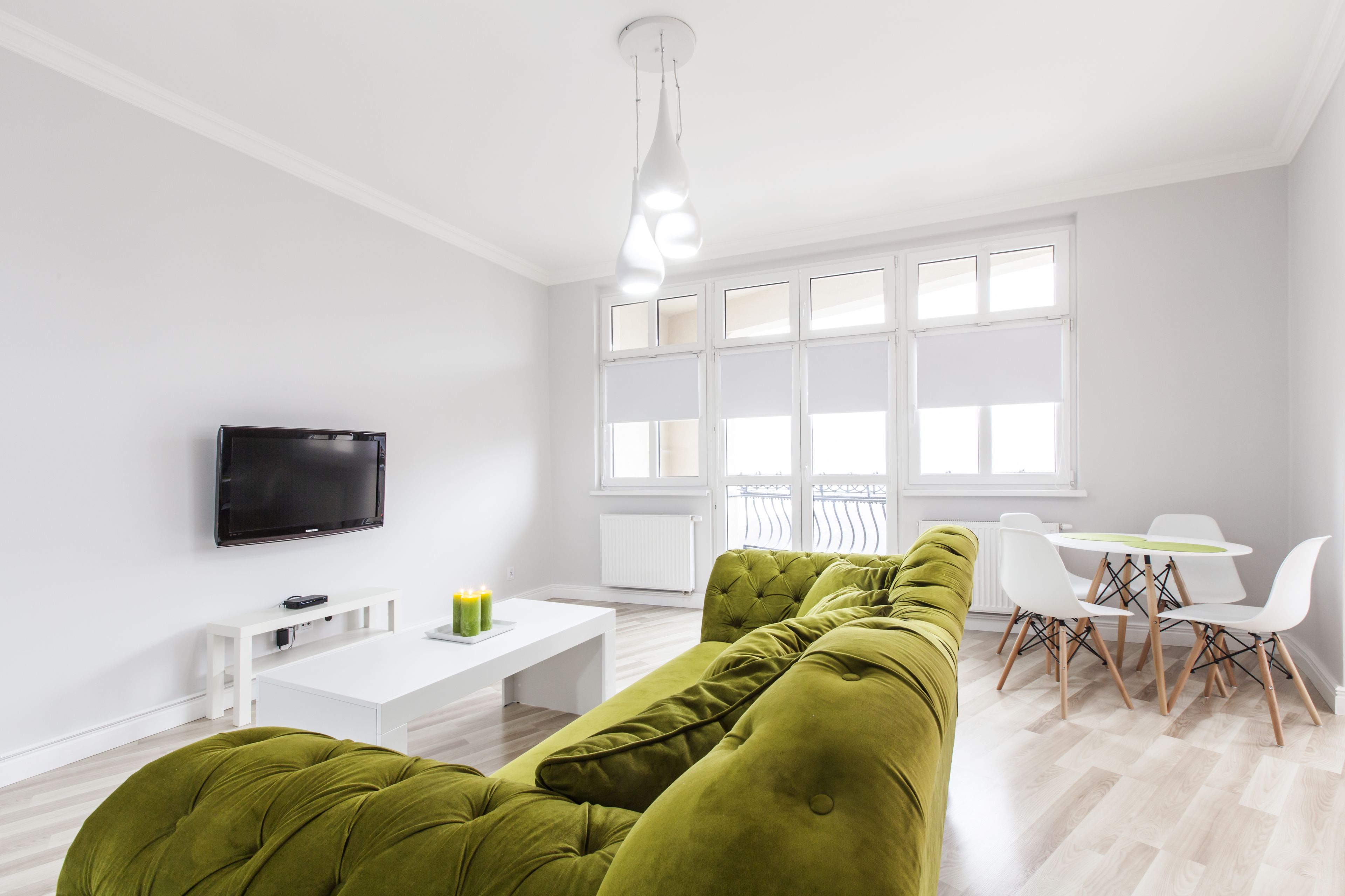 A modern living room with a green sofa, a white coffee table, a flat-screen TV, and a dining area with white chairs and a table.