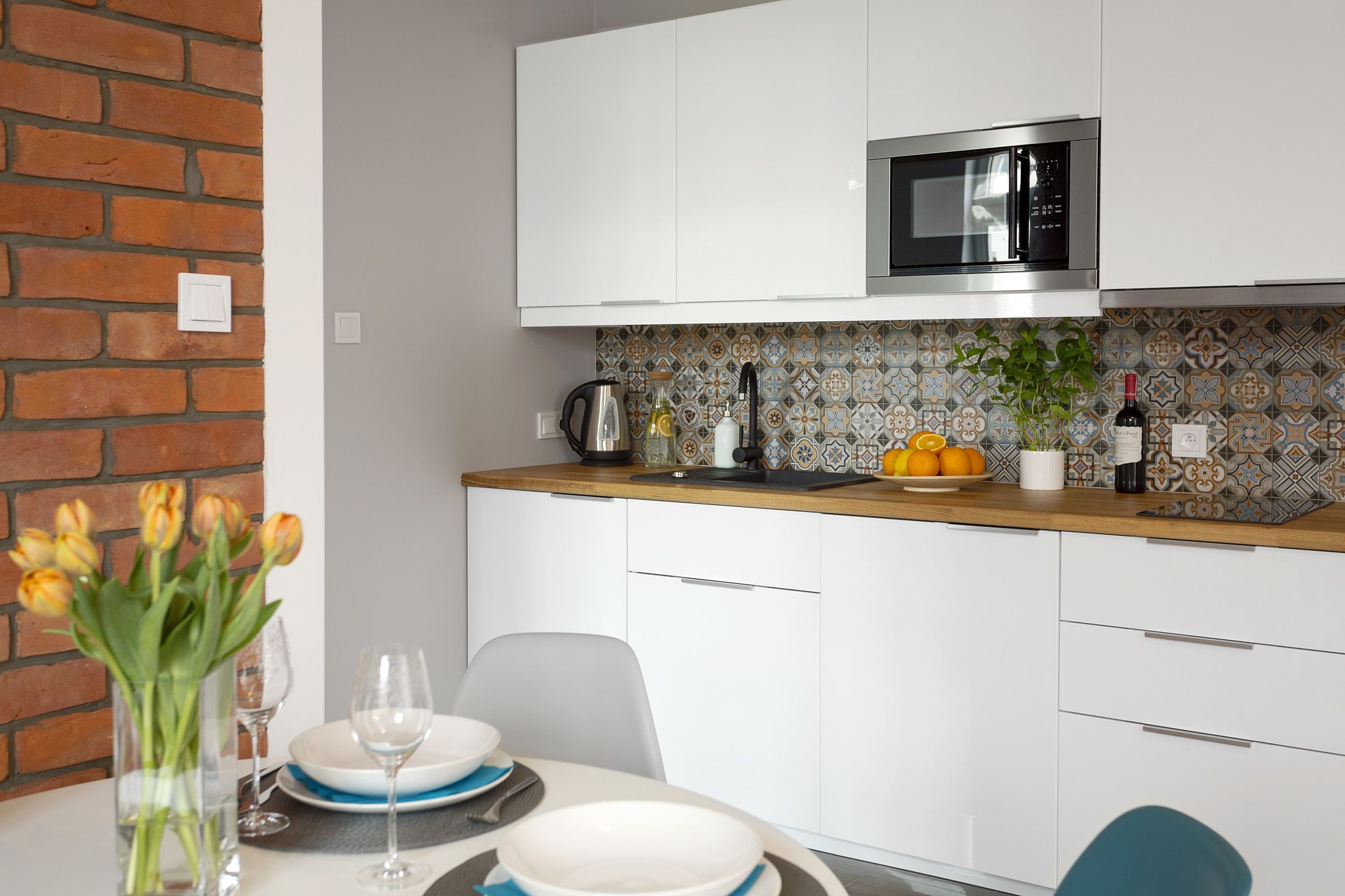 Cozinha moderna com armários brancos, micro-ondas e painel de azulejos. Há uma mesa de jantar com pratos e um vaso de flores.