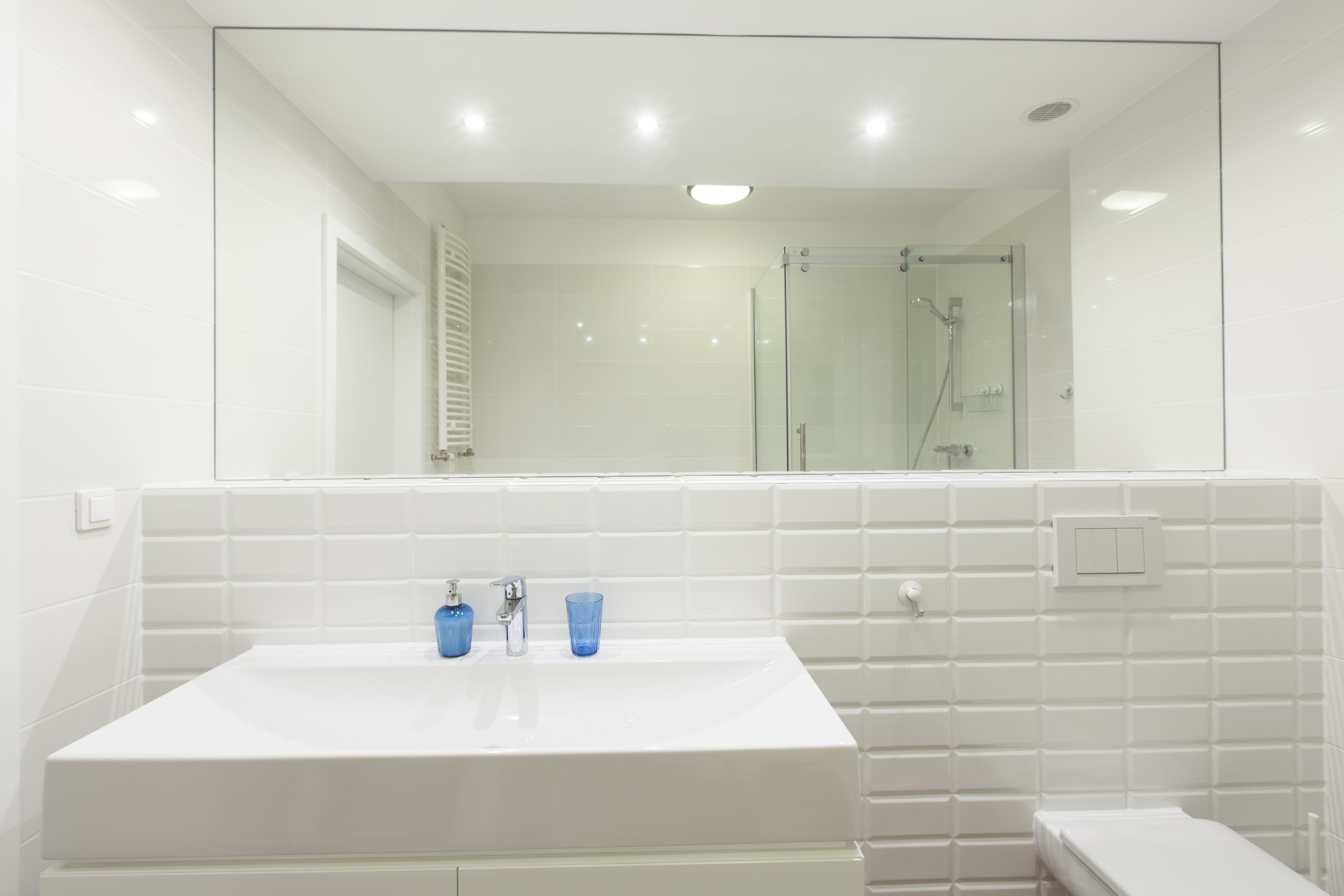 A modern bathroom with a large mirror, white tiles, a sink with two blue glasses, and a glass-enclosed shower.