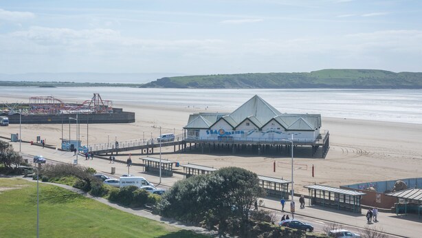 Un edificio costero con un techo destacado y un muelle que se adentra en el mar, junto a una playa arenosa.
