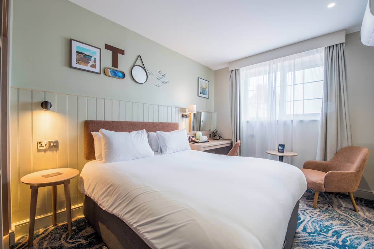 Una habitación de hotel con una cama grande, un escritorio, una silla y un ventanal con cortinas.