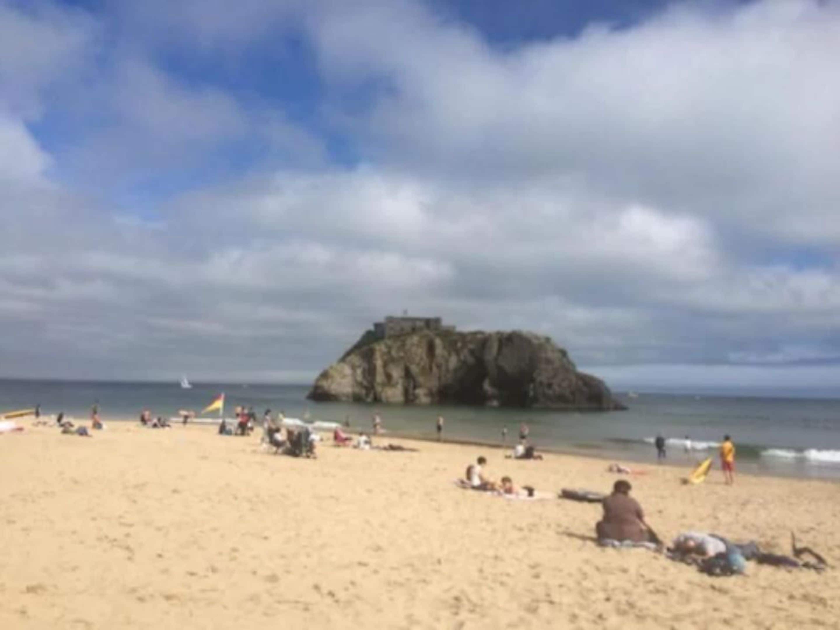 A beach with people sunbathing and swimming, a rocky outcrop in the background, and a clear sky.