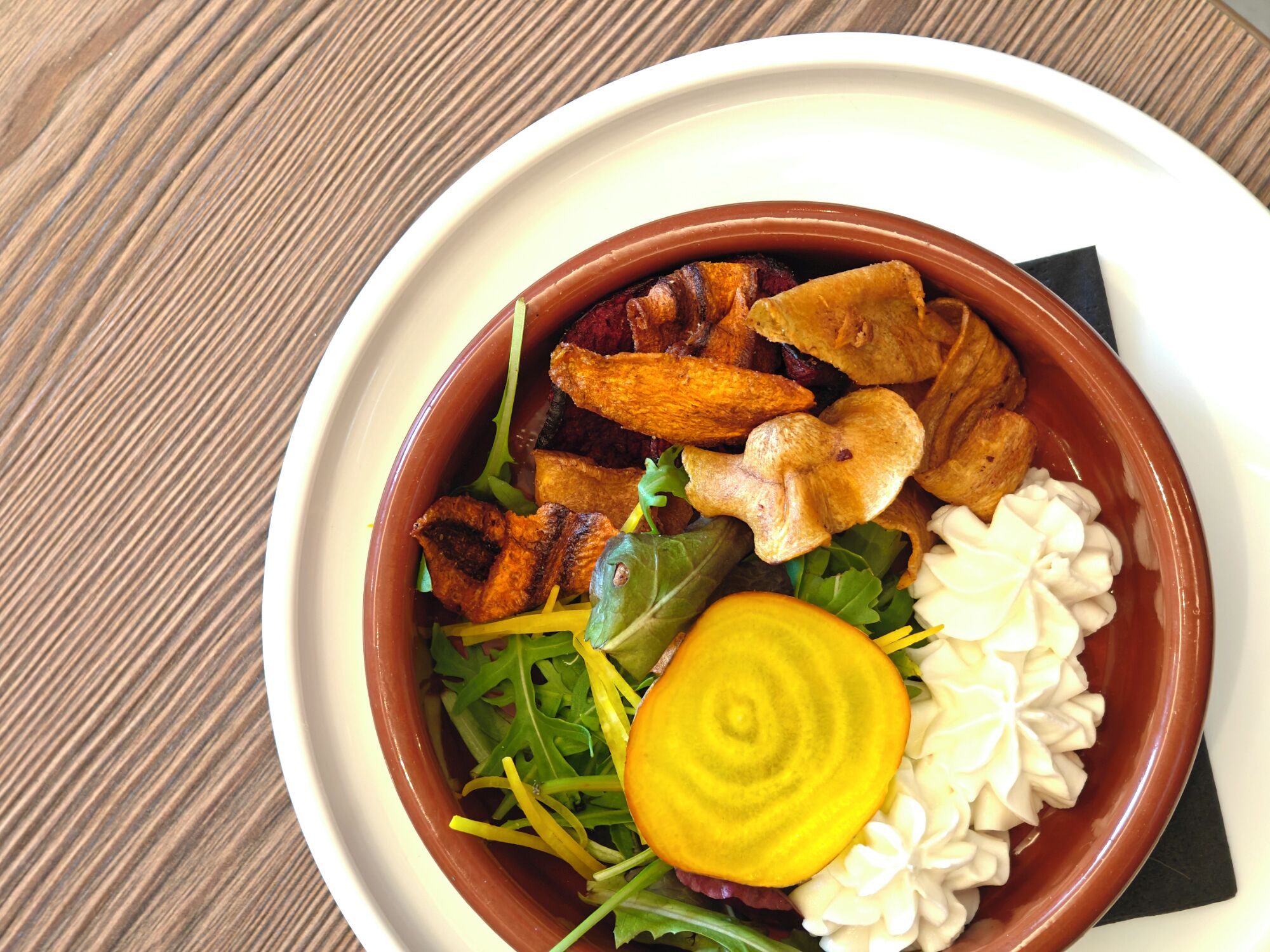 A bowl of food with various items including greens, a slice of beetroot, and some meat.