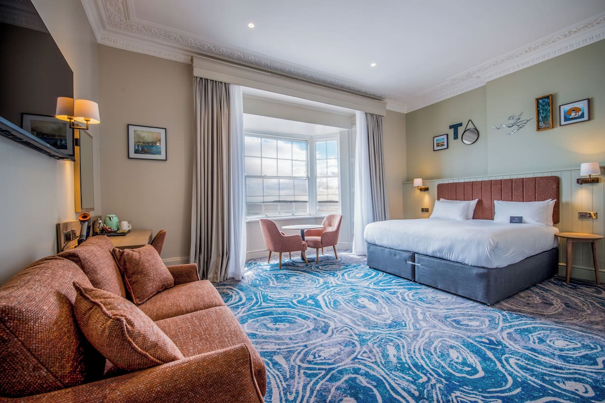 Una habitación de hotel con una cama grande, un sofá, una mesita y una ventana con cortinas.