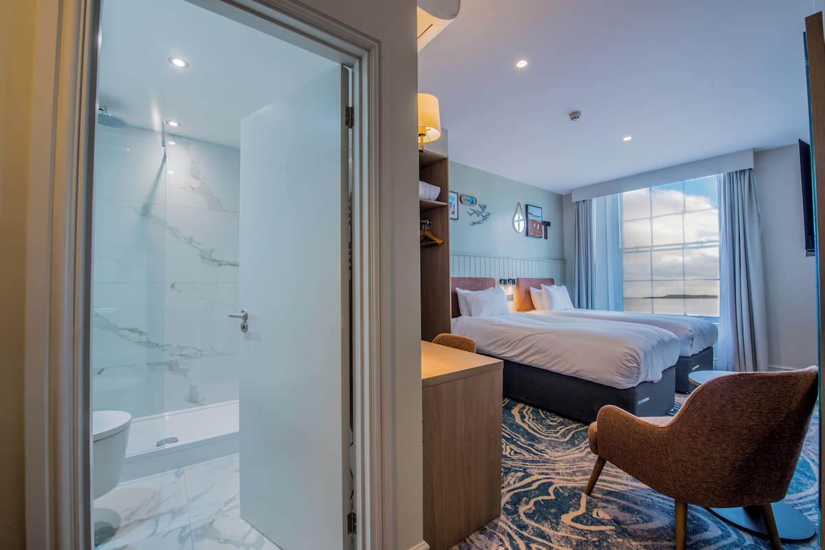 Habitación de hotel con cabina de ducha de vidrio, cama con ropa blanca, escritorio de madera y alfombra estampada.