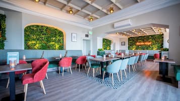 Un restaurante moderno con una mesa común alargada, sillas coloridas y una pared verde con plantas.