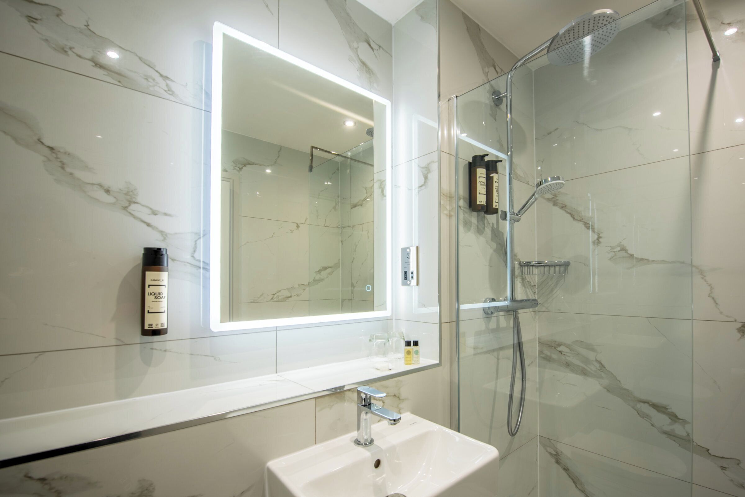 Un baño moderno con un revestimiento de mármol en la pared, un espejo grande, un lavamanos y una zona de ducha con mampara de vidrio.