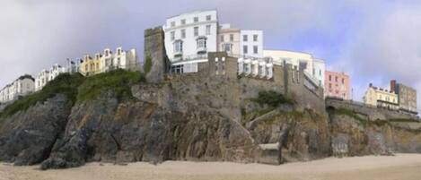 A coastal building with multiple stories, situated on a rocky cliff overlooking a sandy beach.