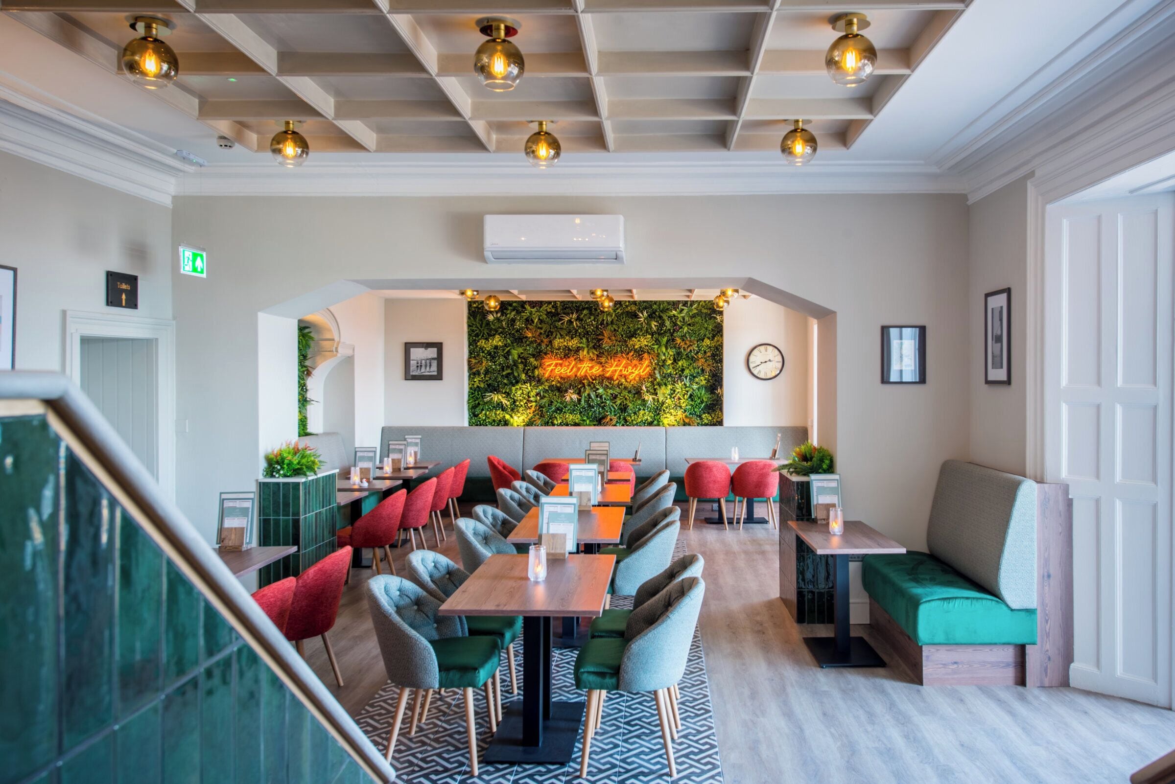 Un restaurant moderne avec une longue table conviviale, des chaises rouges et vertes, et un mur vert orné d’une enseigne indiquant « Grillé à la minute ».