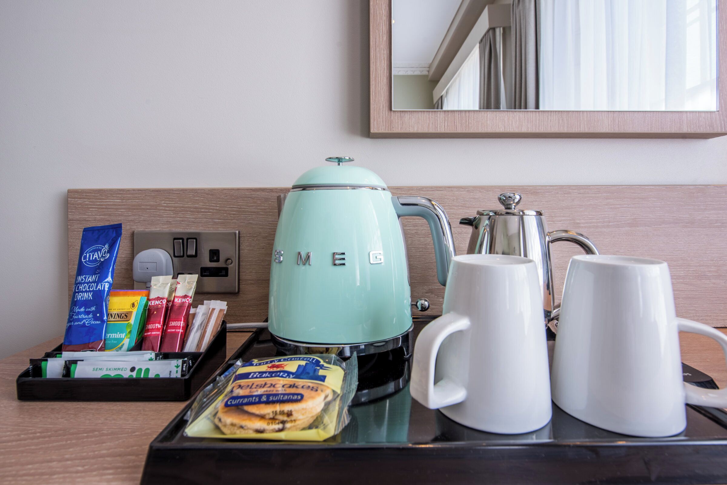A hotel room's breakfast setup with a coffee maker, two white mugs, a tray with snacks, and a mirror on the wall.
