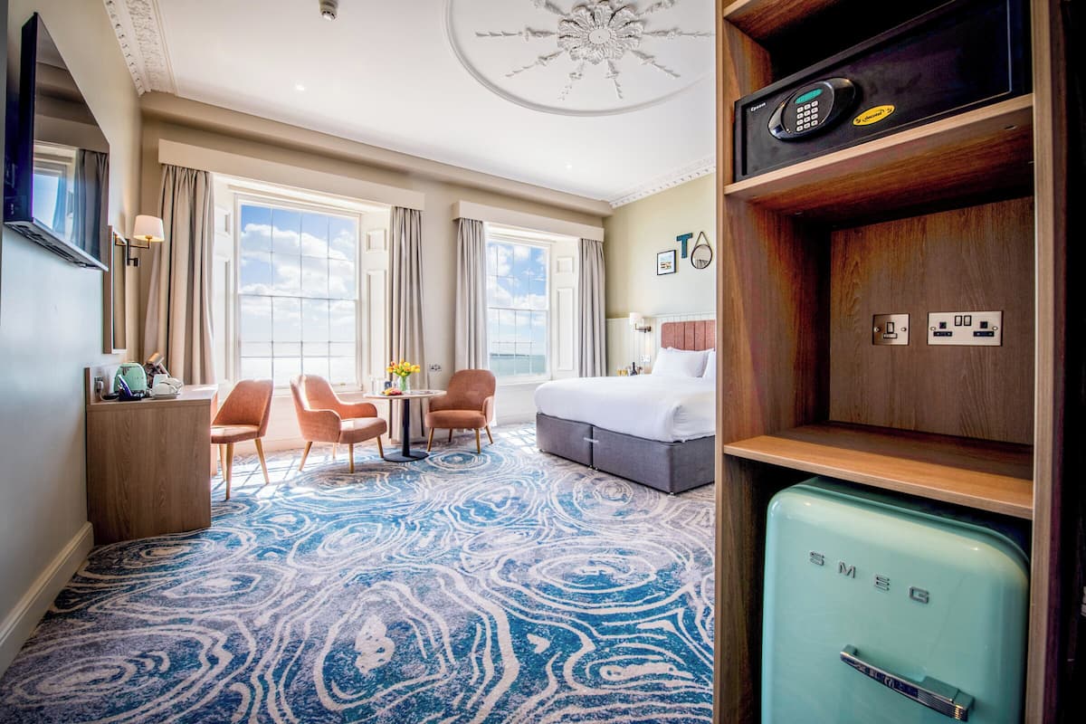 Una habitación de hotel con cama, escritorio, silla, una pequeña mesa con flores, alfombra estampada en azul, refrigerador de estilo vintage y una lámpara en el techo.