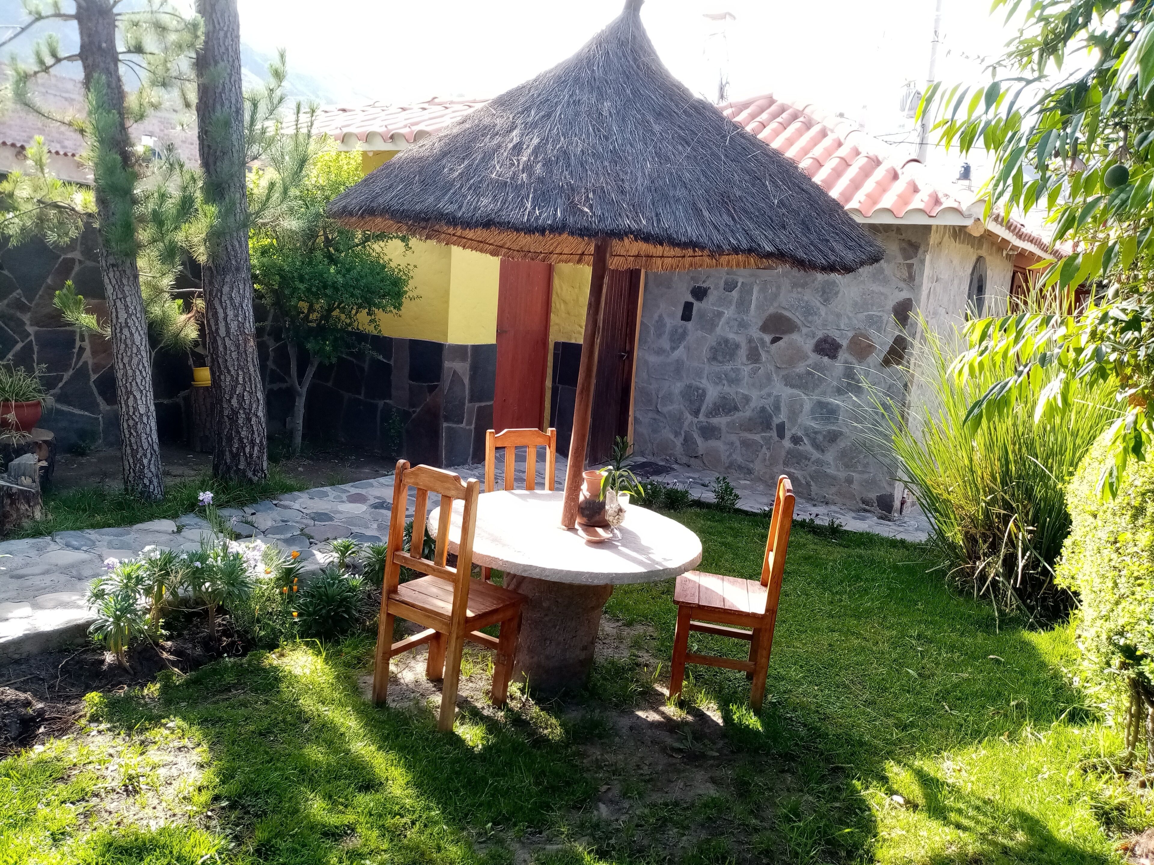 A round wooden table with chairs under a thatched umbrella, surrounded by greenery and a stone wall.