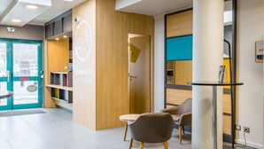 A hotel reception area with a wooden reception desk, a round table, and two chairs. - B&B HOTEL Tours Nord 2 La petit Arche (Tours)