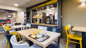 A modern hotel dining area with multiple tables and chairs, a breakfast tray with toast, coffee, and yogurt, and a visible B&B logo. - B&B HOTEL Tours Nord 2 La petit Arche (Tours)