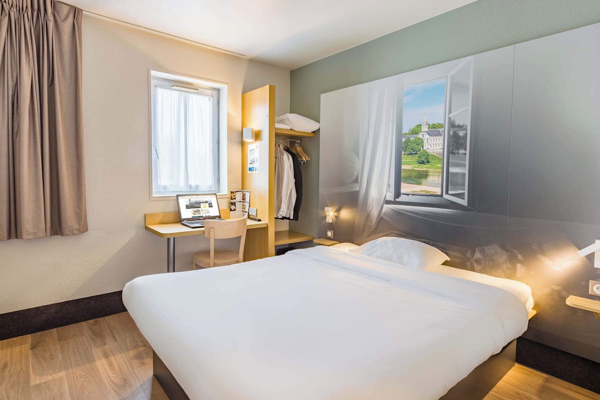 Quarto de hotel com cama, escrivaninha, cadeira, guarda-roupa e janela com vista.
