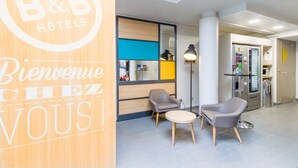 A B&B hotel reception area with a wooden reception desk, two armchairs, a small round table, and a modern interior design. - B&B HOTEL Tours Nord 2 La petit Arche (Tours)