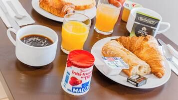 Una escena de desayuno con croissants, un vaso de jugo de naranja, una taza de café, un frasco de mermelada de fresa, una bolsita de té y un cartón de leche sobre una mesa.