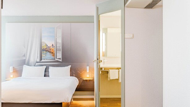 A hotel room with a bed, a painting of a cityscape, a bathroom with a mirror, and a wooden floor. - B&B HOTEL Chatellerault (Chatellerault)