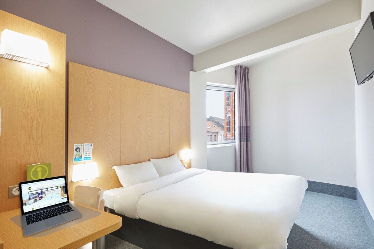 A hotel room with a bed, a desk with a laptop, a bedside lamp, a window with curtains, and a TV mounted on the wall.