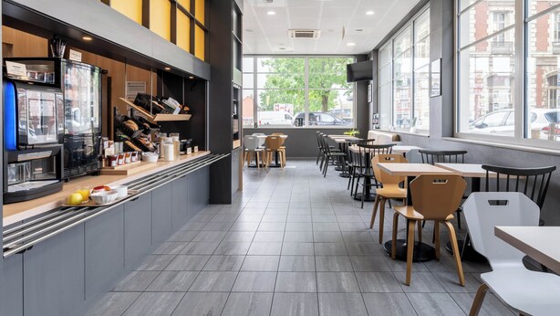 A modern cafe with a counter, seating area, and large windows. - B&B HOTEL Lille Roubaix Campus Gare (Roubaix)