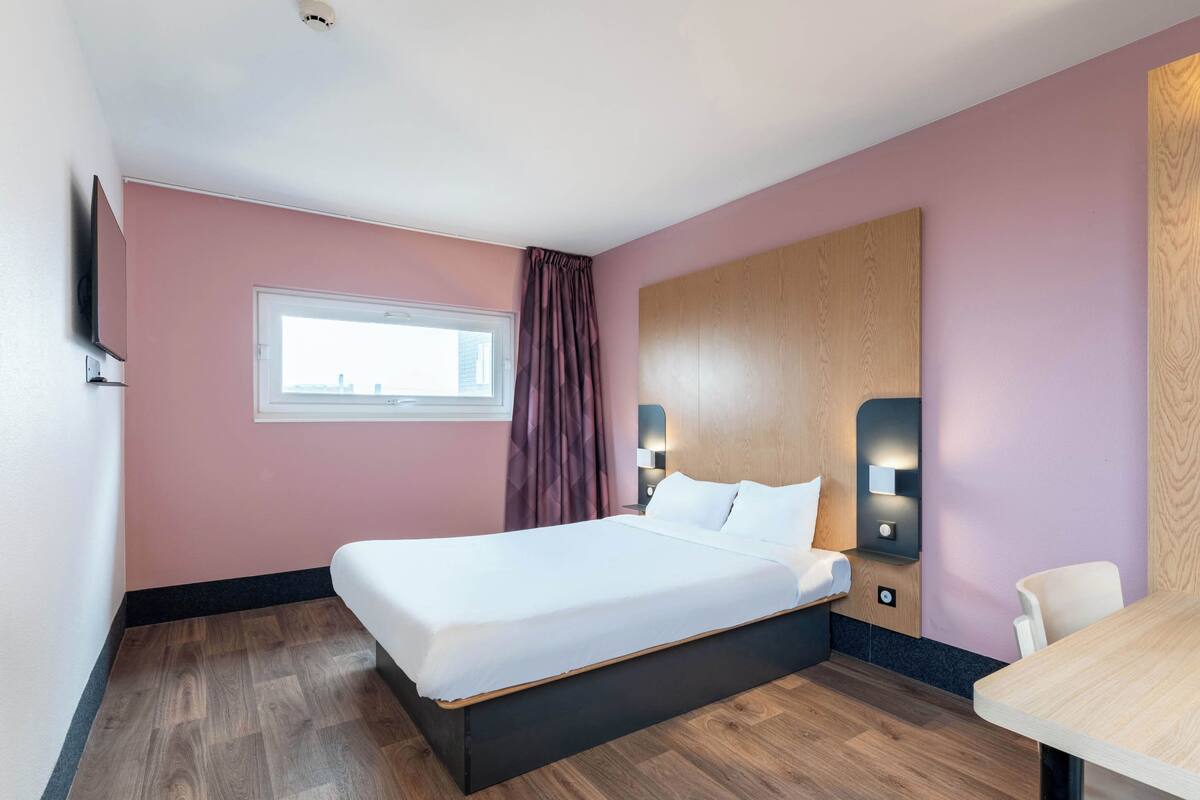 A hotel room with a bed, a desk, a window with curtains, and a wall-mounted TV.