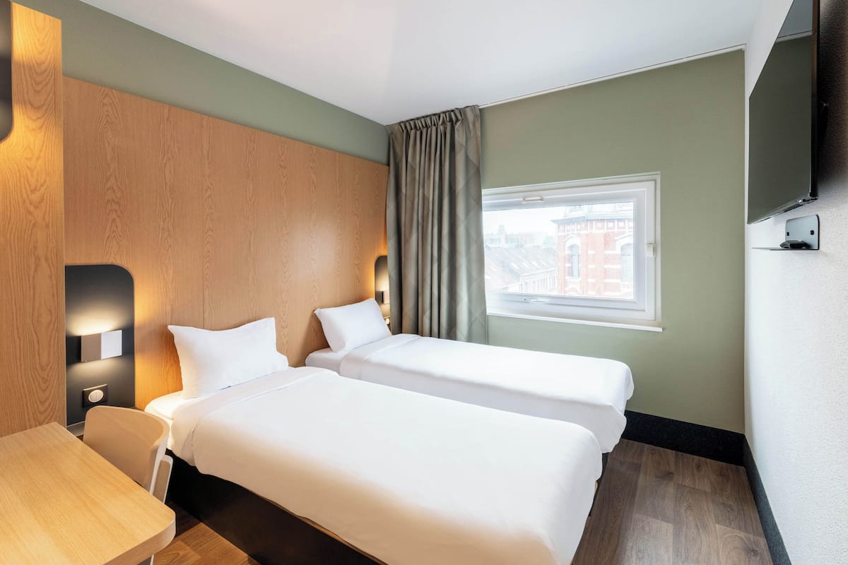 A hotel room with two beds, a wooden headboard, a desk, a chair, a TV, and a window with curtains.