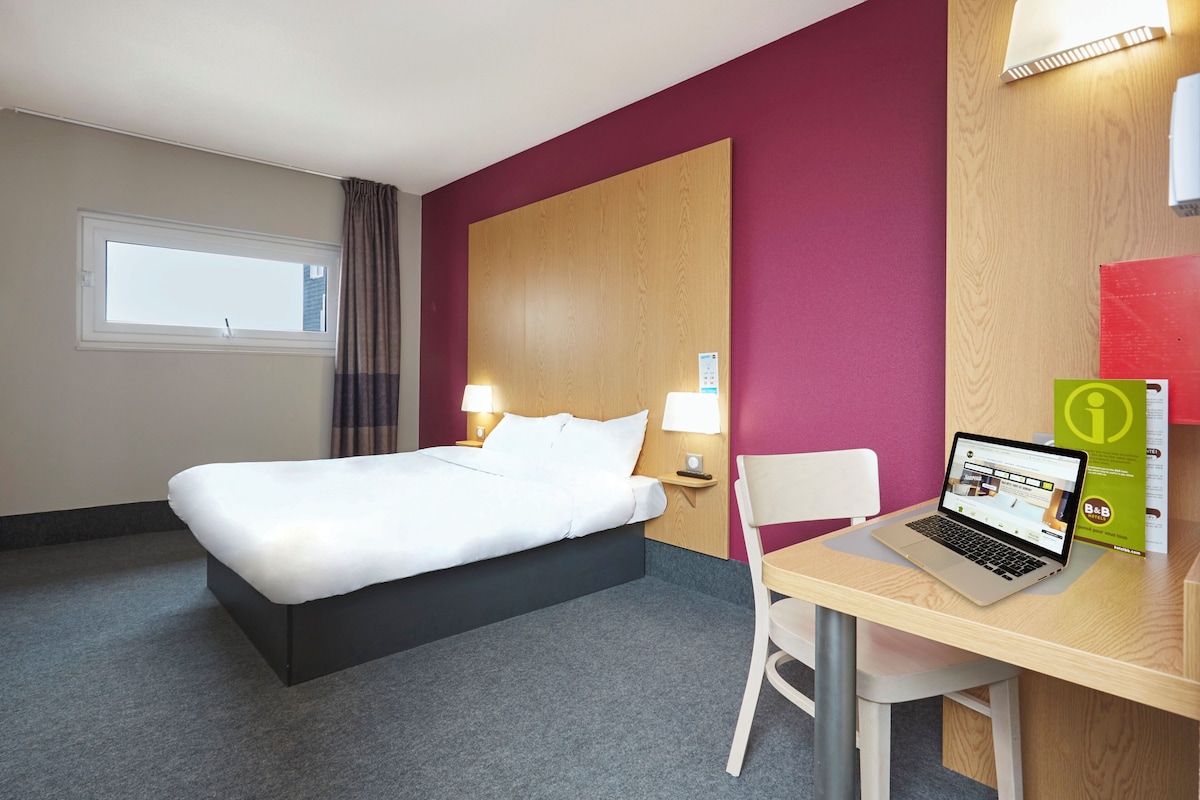 A hotel room with a bed, a desk with a laptop, a chair, a window, and a wall-mounted lamp.