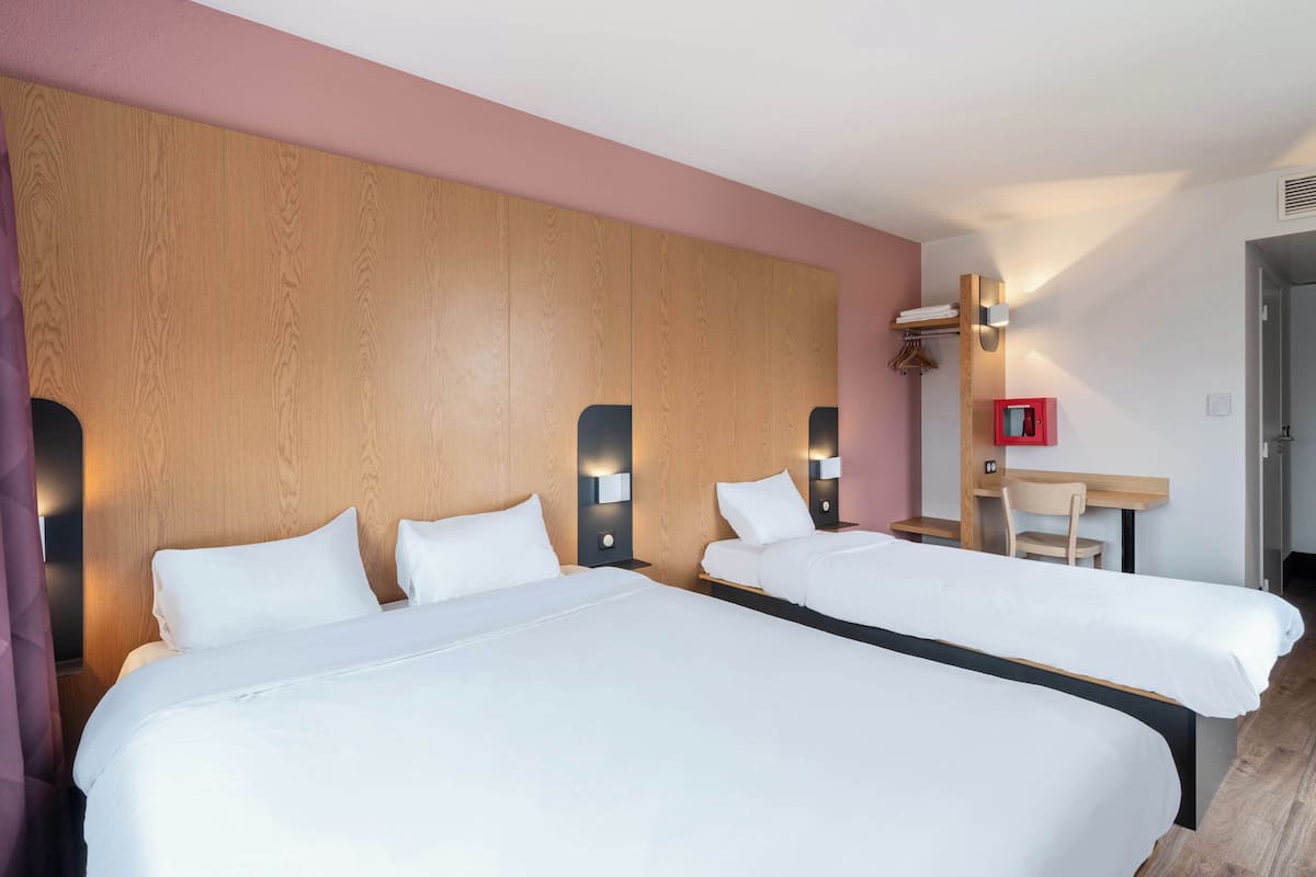 A hotel room with two beds, wooden headboard, bedside tables with lamps, a desk, and a red emergency box.