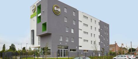 A modern B&B hotel with a grey and white facade, multiple windows, and a parking lot with cars.