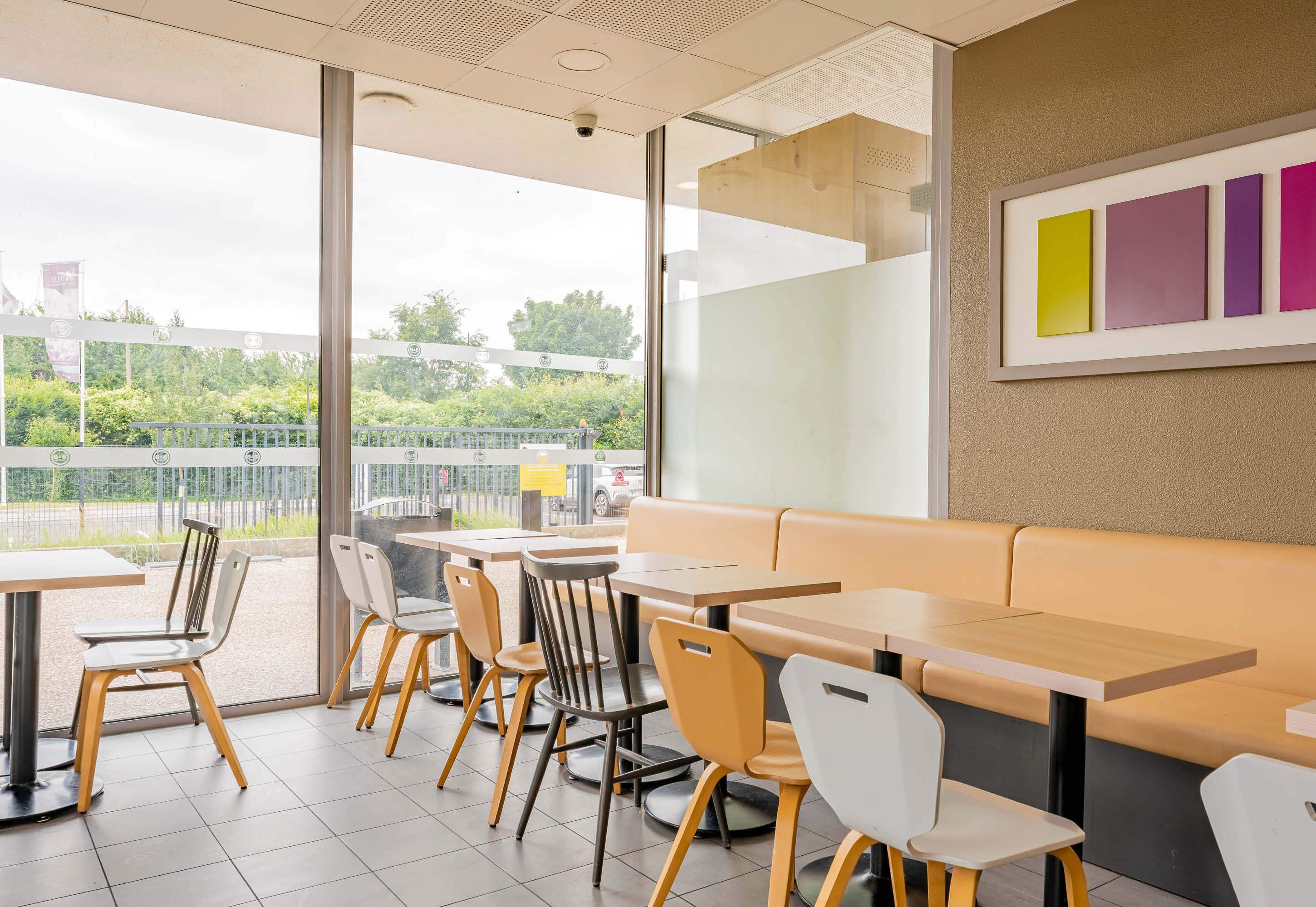 A modern cafe with a seating area featuring tables and chairs, a large window with a view of greenery and a road, and a wall with a framed artwork.