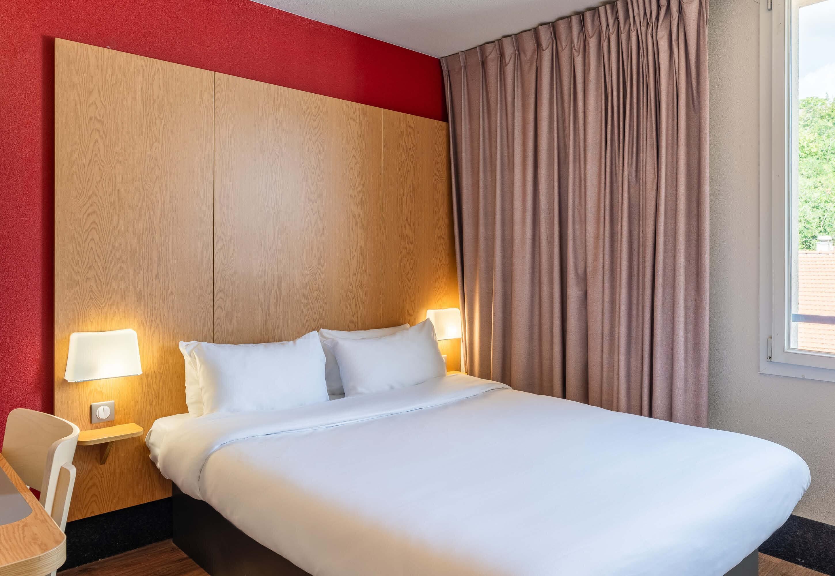 Ein Hotelzimmer mit einem großen Bett, zwei Nachttischlampen, einem Holzkopfteil, einem Fenster mit Vorhängen und einem kleinen Tisch mit einem Stuhl.
