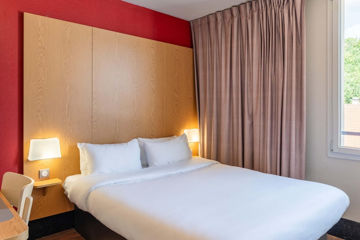 A hotel room with a large bed, two bedside lamps, a wooden headboard, a window with curtains, and a small table with a chair.